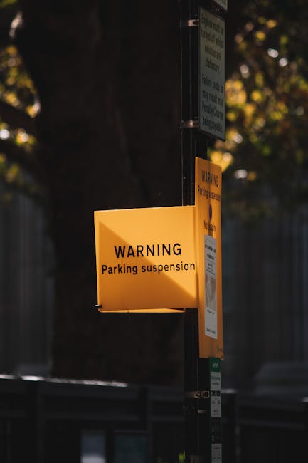 A yellow warning sign attached to a metal pole, displaying the message 'WARNING Parking suspension' in black text, situated outdoors during daylight hours. The sign is illuminated by natural light, with a background of blurred trees and a fence. This signage indicates a temporary parking restriction relevant to house removals and moving logistics in the RM14 area, as managed by Man with Van Upminster Bridge. The sign’s visibility and clear messaging support the planning and execution of home relocation services, particularly in managing parking suspensions during furniture transport and loading processes for local house moves.