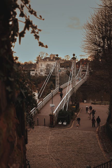 A street scene during daylight with a view of a white suspension bridge extending over a river or valley, supported by vertical cables and towers. The background features a large, stately building with multiple windows, surrounded by trees with bare branches, indicating a colder season. On the cobbled street below, several pedestrians are walking, some carrying bags or backpacks, with a few groups in conversation. Near the bridge, a man wearing dark clothing is seen walking along the pavement, which is bordered by a low wall and a small iron fence. The scene is illuminated by natural light, casting soft shadows, and the overall setting suggests an urban area with historical or architectural significance, relevant to home relocations or furniture transport services provided by companies like Man with Van Upminster Bridge.