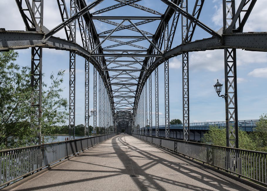 A view of a steel truss bridge with intricate lattice structures overhead, spanning over a river on a bright, partly cloudy day. The bridge's walkway is paved with asphalt, lined on both sides with metal railings. On the right side, there are two traditional-style street lamps attached to the bridge's framework, and on the left, leafy green trees extend into the frame. Shadows of the metal lattice are cast onto the pathway, creating a pattern of light and dark. This scene captures the structural complexity of the bridge designed for pedestrians and possibly light vehicle traffic, emphasizing the metallic construction and open sky above. The location appears to be part of a scenic route that could facilitate home relocation or furniture transport, as offered by professional removal services like Man with Van Upminster Bridge, associated with the page 'Best Removal Routes from Upminster Bridge Station to RM14 Homes, UPMINSTER BRIDGE.'