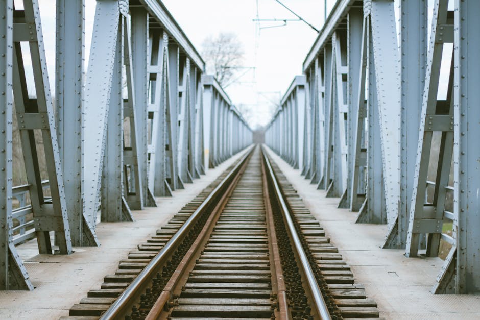 A view of a railway bridge with metal trusses painted in light grey, supporting two metal railway tracks running through the middle. The tracks are flanked by wooden sleepers and gravel, extending into the distance with a vanishing point at the horizon. Overhead, there are electrical wires and supporting pylons. The scene is outdoors, with natural daylight illuminating the structure. This infrastructure is part of the transportation routes often utilized during home relocation and furniture transport, and the image is relevant for illustrating the logistics involved in packing, moving, and loading processes that [COMPANY_NAME], such as Man with Van Upminster Bridge, facilitates in residential removals.