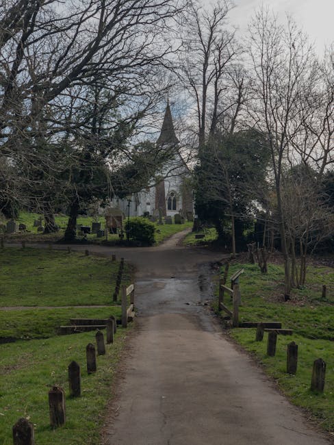 A paved pathway with wooden bollards on either side, leading through a green area with grass and sparse trees towards a white church with a pointed steeple in the background. The scene is outdoors under an overcast sky, with leafless branches revealing the church's architecture. This setting reflects the outdoor environment involved in house removals and furniture transport, highlighting the importance of careful logistical planning in home relocation services by Man with Van Upminster Bridge.