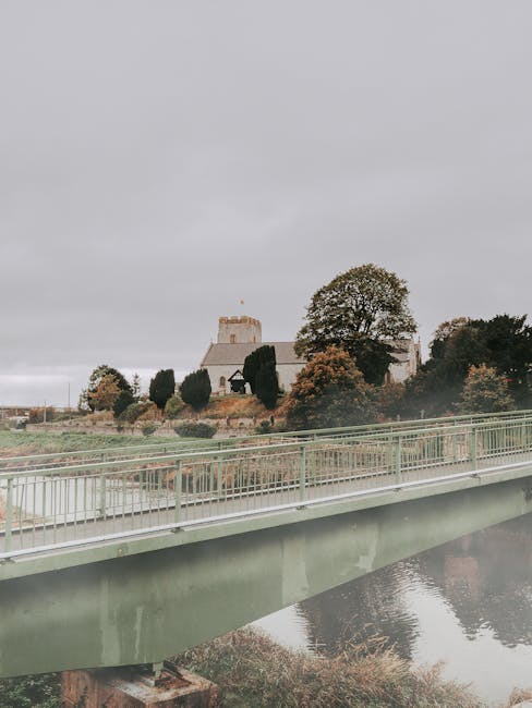 The image shows a view of a historic castle situated on a hill, surrounded by trees with a mix of green and autumnal leaves. In the foreground, there is a modern light green bridge with metal railings crossing over a calm river, reflecting the bridge and nearby foliage. The sky is overcast with gray clouds, creating a subdued lighting environment. Inside the scene, partially visible on the right side, are stacked cardboard boxes and wrapped furniture, indicating a home relocation process. The boxes are placed on a pavement near the bridge, while the furniture, covered in protective fabric or plastic, is either inside or near a moving van parked close to the property. The setting suggests a professional removals operation by [COMPANY_NAME], with the process of loading household items into a vehicle for transport, highlighting the logistical aspects of furniture transport and packing during a house move from Cranham Road to St Mary's Lane in UPMINSTER BRIDGE.