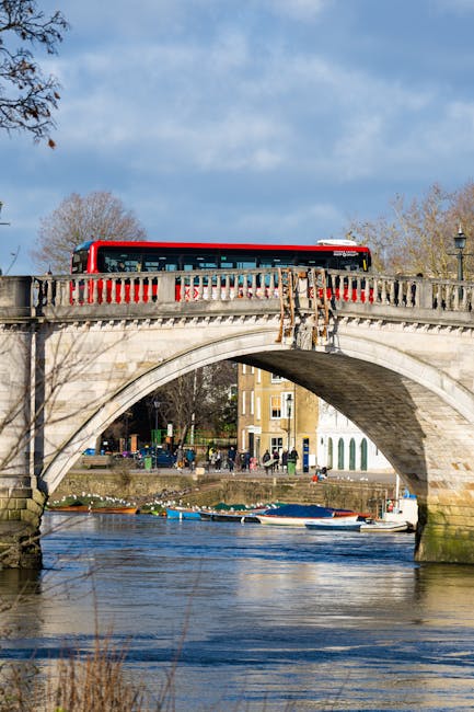 A red and white bus is positioned on the top of a stone arch bridge over a river in Upminster Bridge, with the bus stopping or passing across the bridge. The bridge features a detailed balustrade with vertical stone balusters and decorative elements. Below, the river flows calmly, with several small boats docked along the water's edge. Behind the bridge, trees with sparse autumn foliage and a row of residential buildings are visible, and the sky above is partly cloudy with patches of blue. The scene depicts a typical urban environment suitable for house removals and logistics, with the bridge serving as a key transit point in the streetscape, supported by a context of residential and recreational areas. This setting aligns with the process of furniture transport and home relocation in the Upminster Bridge area, where careful movement of household items often involves crossing bridges and navigating urban waterways. Man with Van Upminster Bridge, operating in the RM14 and RM11 postcode areas, provides services related to removals and moving logistics in such environments.