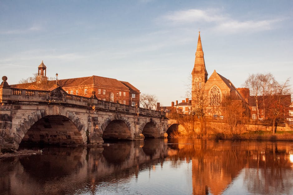 A stone bridge with multiple arches spans over a calm river in Upminster Bridge, with a historic church featuring a tall spire and a large stained-glass window visible in the background. The bridge is lined with a stone balustrade, and several buildings with brick facades are situated behind it. The scene is illuminated by warm, natural light from the setting or rising sun, casting a golden glow on the buildings and their reflections in the water. Leafless trees are present near the church, and a clear sky with soft clouds forms the backdrop. This image captures the tranquil environment typical of a house relocation or furniture transport setting in a historic townscape, illustrating the architectural and scenic elements involved in a move facilitated by professional removals services like Man with Van Upminster Bridge.