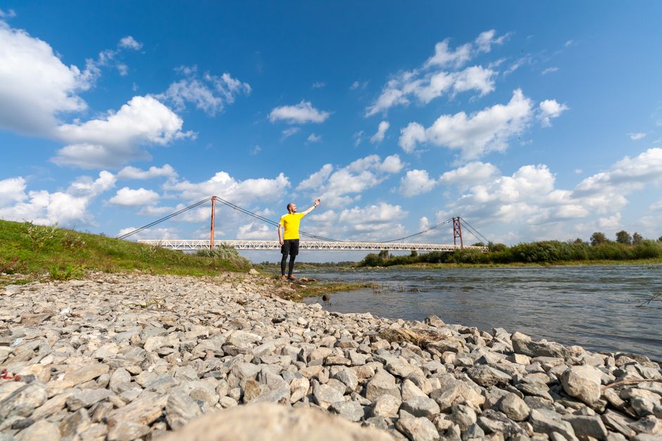 A man wearing a yellow shirt and dark pants stands on a rocky riverside shoreline, holding a fishing rod with the line cast into the water. In the background, there is a large suspension bridge spanning the river, with a blue sky filled with scattered white clouds overhead. The scene is outdoors, featuring natural elements such as the river, greenery on the riverbank, and the open sky, illustrating a tranquil setting. This image relates to home relocation and packing and moving processes, with [COMPANY_NAME] providing professional removals services, including waste disposal after an Upminster Bridge clear-out, as referenced on the webpage handling waste disposal after an Upminster Bridge clear-out, UPMINSTER BRIDGE.