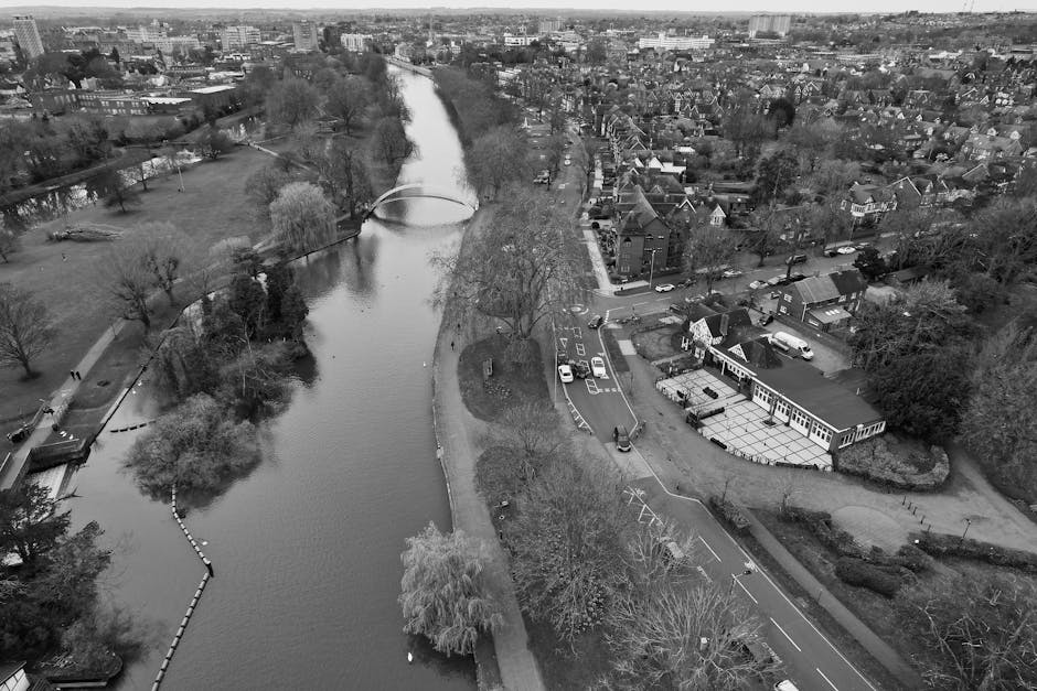 Aerial black-and-white photograph showing a river winding through a residential area with trees along its banks and a small bridge crossing over the water. On the right side, there is a parking area with several cars parked, adjacent to a single-story building with large windows, which appears to be a commercial or community space. The surrounding neighborhood consists of numerous houses with pitched roofs, trees, and roads, some of which are empty of traffic. The scene captures the area where house removals and furniture transport might take place, with visible open spaces and access roads. The overall environment appears calm and organized, emphasizing logistics for house removals or moving services managed by [COMPANY_NAME], especially considering the parking and loading zones near the property.