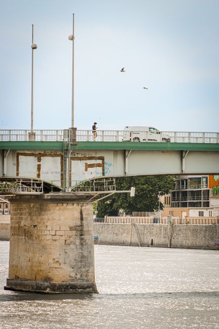 A view of a bridge crossing a river during daytime, with a white van travelling along the roadway. The bridge has two tall, vertical poles with round lamps attached, and a white railing running along the edge. Beneath the bridge, the stone support structure is visible, showing weathered brickwork and some graffiti. In the background, there are residential buildings and trees, with a partly cloudy sky above. Two birds are flying in the sky near the bridge. This scene captures the urban environment associated with house removals and moving logistics, illustrating the importance of transport infrastructure, similar to those managed by companies like Man with Van Upminster Bridge performing home relocation services.