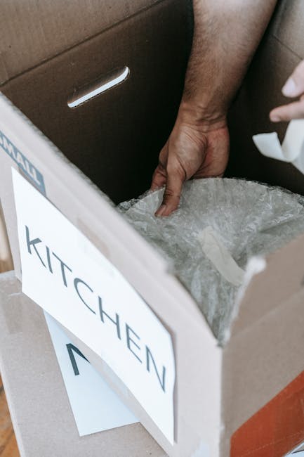 Photograph showing a person's hand grasping the top of a cardboard box labeled 'KITCHEN' with a printed sign, situated inside a vehicle or on a loading area. The box is filled with packing material, likely bubble wrap or paper, to protect contents during a house removal. The box is positioned next to a metallic trolley or dolly, which is partially visible in the foreground, indicating an active loading process. In the background, part of a cardboard interior wall or partition is visible, suggesting the scene is set within a moving van or a storage space used during relocation. The lighting is natural, highlighting the textures of the cardboard, packing material, and the person's hand, emphasizing the careful handling involved in packing and transportation during home relocation, as facilitated by Man with Van Upminster Bridge.