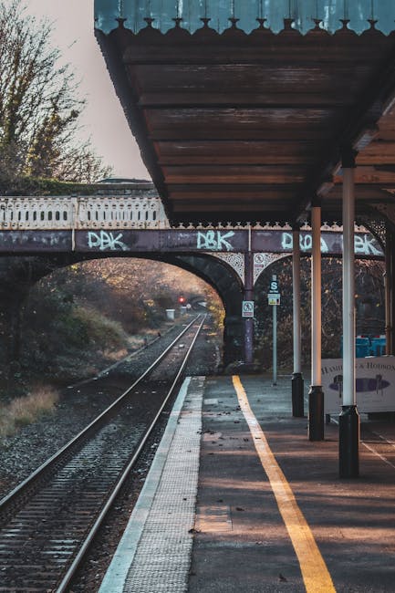 A view of an outdoor train station platform with a covered waiting area supported by white metal poles. The platform has a yellow safety line along the edge and is paved with concrete. Overhead, a dark-colored roof extends over the platform, providing shelter for passengers. In the background, railway tracks run straight into the distance, with a bridge crossing over the tracks featuring graffiti-covered walls and a stone arch. The surrounding landscape includes trees visible beyond the tracks, and a signal light is visible further along the line. This scene depicts a typical station setting suitable for home relocation or furniture transport, as provided by Man with Van Upminster Bridge, supporting efficient moving logistics and packing preparations for residents on Station Road and Upminster Bridge.