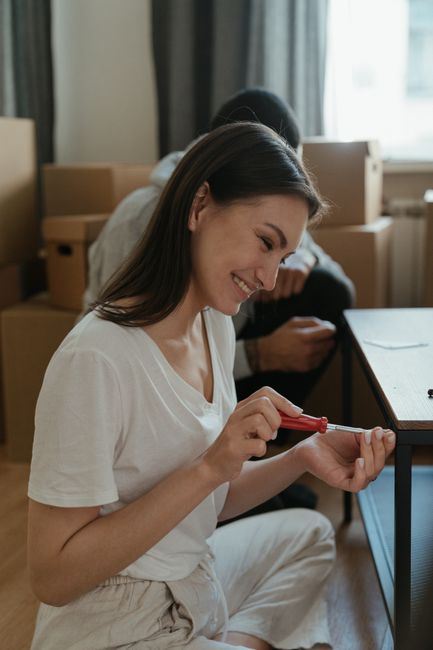 A woman with long dark hair, wearing a white t-shirt and light-colored trousers, sits on the floor inside a room during a home relocation. She is smiling while using a red-handled screwdriver to assemble or disassemble a piece of furniture, possibly a small table or desk. Behind her, there are cardboard boxes stacked on the floor, indicating packing and moving preparations. A window with curtains provides natural light, illuminating the scene. The room has a neutral colour scheme with a wooden floor and simple furnishings. This image illustrates the process of packing, furniture assembly, or disassembly as part of a house removal service, reflecting the careful handling and logistical planning undertaken by professionals like Man with Van Upminster Bridge during a house move.