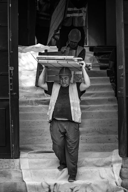 A professional mover from Man with Van Upminster Bridge is transferring a large wooden dining table, which is wrapped in plastic for protection, down a short flight of steps at a residential property situated on steep St Mary's Lane in UPMINSTER BRIDGE. The mover, wearing a high-visibility vest and casual trousers, is carefully balancing the table on his shoulders while walking on a protective sheet laid across the steps. Behind him, another staff member is seen carrying a cardboard box wrapped in packing paper, and a van is parked nearby, likely for loading the furniture and belongings for home relocation or furniture transport. The environment suggests a coordinated loading process typical of removals services, with an emphasis on safe handling of heavy household furniture during a house move.