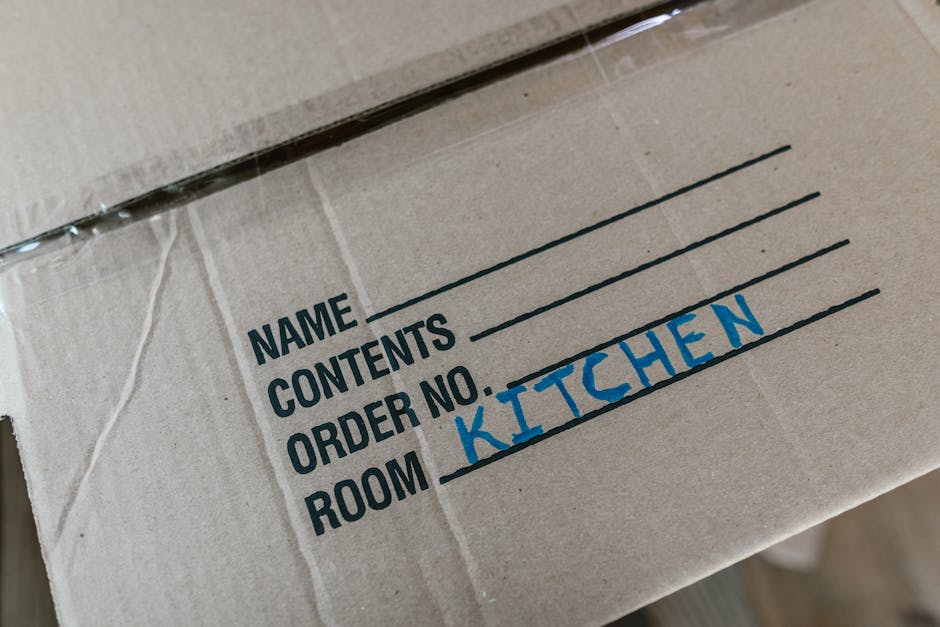 A woman with long dark hair, wearing a white t-shirt and light-colored trousers, sits on the floor inside a room during a home relocation. She is smiling while using a red-handled screwdriver to assemble or disassemble a piece of furniture, possibly a small table or desk. Behind her, there are cardboard boxes stacked on the floor, indicating packing and moving preparations. A window with curtains provides natural light, illuminating the scene. The room has a neutral colour scheme with a wooden floor and simple furnishings. This image illustrates the process of packing, furniture assembly, or disassembly as part of a house removal service, reflecting the careful handling and logistical planning undertaken by professionals like Man with Van Upminster Bridge during a house move.