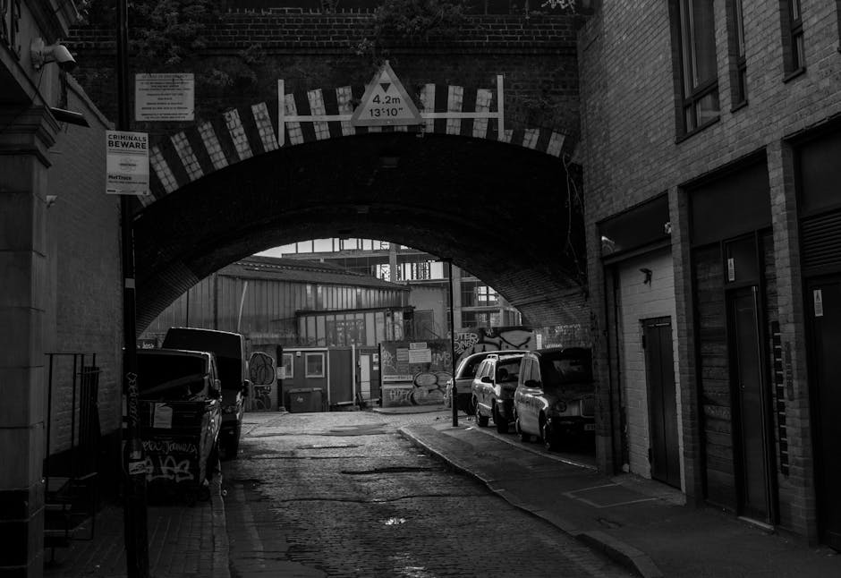 A wide-angle view of a narrow street under a cloudy sky in Upminster Bridge, showing a decorative cast-iron bridge with a red and yellow railing arching over a small waterway. On the pavement beside the bridge, a black van is parked, and a tall black and yellow traffic sign is visible on the right side of the image. In the background, several people are gathered near the entrance to a building, possibly engaged in a house removal or furniture transport activity. Surrounding buildings include a large yellow structure on the left with multiple windows and green plants growing nearby, as well as older brick buildings with varied window styles further along the street. The scene captures the logistics of urban furniture and appliance transportation, with people, vehicles, and equipment indicating a home relocation process managed by Man with Van Upminster Bridge.