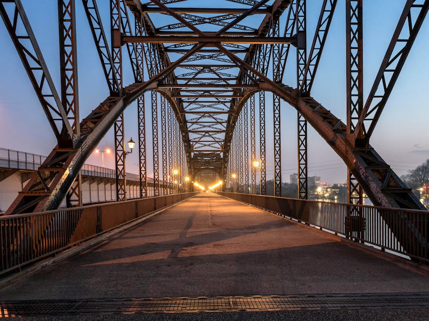 A large steel bridge with intricate lattice framework extending into the distance, illuminated by a row of streetlights along its pedestrian walkway. The structure features arched support beams and vertical trusses, painted in a dark color, contrasting with the soft evening sky transitioning from blue to dusky pink. The walkway appears to be paved with asphalt and is bordered by metal railings. In the background, urban lights are faintly visible, indicating nearby buildings and cityscape. The bridge's setting and construction details suggest it is used for pedestrian or light vehicle access, potentially part of a home relocation or moving process where larger vehicles like vans could load and unload nearby. This image is associated with house removals and furniture transport services offered by Man with Van Upminster Bridge, emphasizing the logistics involved in moving house contents across such infrastructure.
